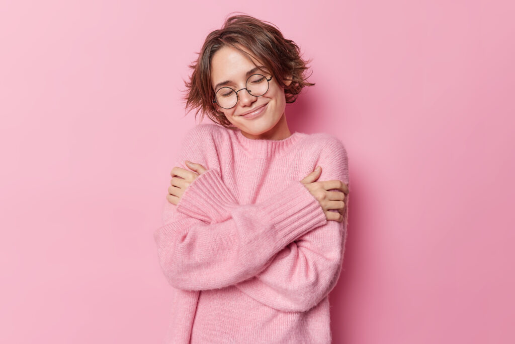 photo tender pretty woman with short hairs embraces herself feels comfortable new soft jumper wears round spectacles vision correction isolated pink background self care concept